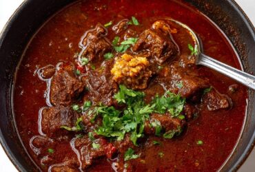Bowl of Texas chili with chunks of beef, deep red broth, melted cheese, sour cream, jalapeños, and cornbread