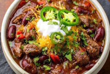 Bowl of spicy steak chili with chunks of seared beef, beans, peppers, melted cheese, sour cream, and cilantro