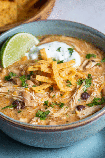 White chicken chili in a rustic bowl with shredded chicken, white beans, green chiles, creamy broth, topped with cheese, cilantro, lime, tortilla strips