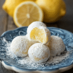 Lemon truffles on a marble surface dusted with powdered sugar, pale yellow centers visible, lemon zest scattered around, fresh lemon halves