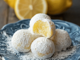 Lemon truffles on a marble surface dusted with powdered sugar, pale yellow centers visible, lemon zest scattered around, fresh lemon halves