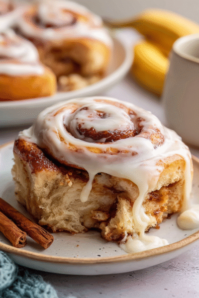 Banana bread cinnamon rolls in a baking dish with golden tops, visible cinnamon-banana swirl, cream cheese glaze drizzled on top