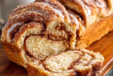 Cinnamon roll bread loaf cut to show soft swirled interior with cinnamon-sugar pattern, cream cheese glaze drizzled on top, golden crust