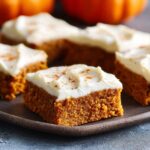Pumpkin bars with cream cheese frosting cut into squares on a wooden board, moist orange pumpkin cake with thick white frosting, cinnamon dust on top