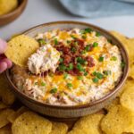 Cheesy bacon ranch dip in a baking dish with golden bubbling cheese top, crispy bacon crumbles, green chive sprinkles, chips and crackers around for dipping