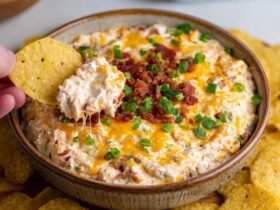 Cheesy bacon ranch dip in a baking dish with golden bubbling cheese top, crispy bacon crumbles, green chive sprinkles, chips and crackers around for dipping