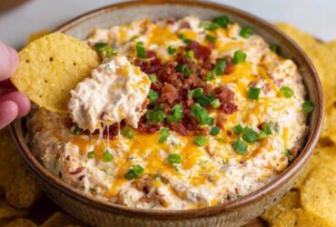 Cheesy bacon ranch dip in a baking dish with golden bubbling cheese top, crispy bacon crumbles, green chive sprinkles, chips and crackers around for dipping