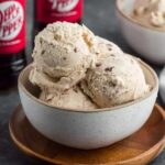 Dr Pepper ice cream scooped into a waffle cone with creamy beige-pink color, soda-reduction swirl visible, drip on side, cherry on top, dark soda in glass behind