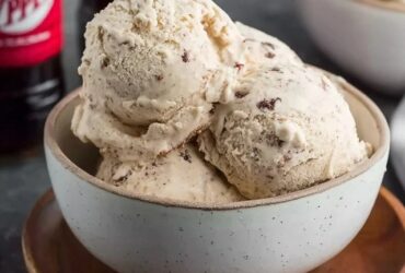 Dr Pepper ice cream scooped into a waffle cone with creamy beige-pink color, soda-reduction swirl visible, drip on side, cherry on top, dark soda in glass behind