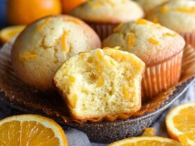 Orange muffins on a wire rack with golden domed tops, orange zest specks visible throughout, soft tender crumb, parchment liners, fresh oranges and zest curls around