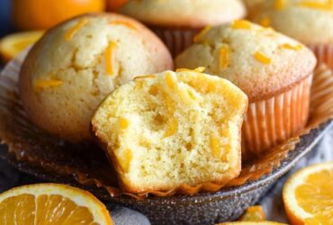 Orange muffins on a wire rack with golden domed tops, orange zest specks visible throughout, soft tender crumb, parchment liners, fresh oranges and zest curls around