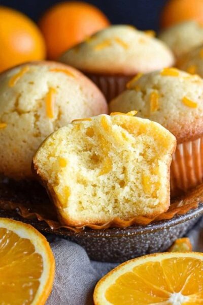 Orange muffins on a wire rack with golden domed tops, orange zest specks visible throughout, soft tender crumb, parchment liners, fresh oranges and zest curls around