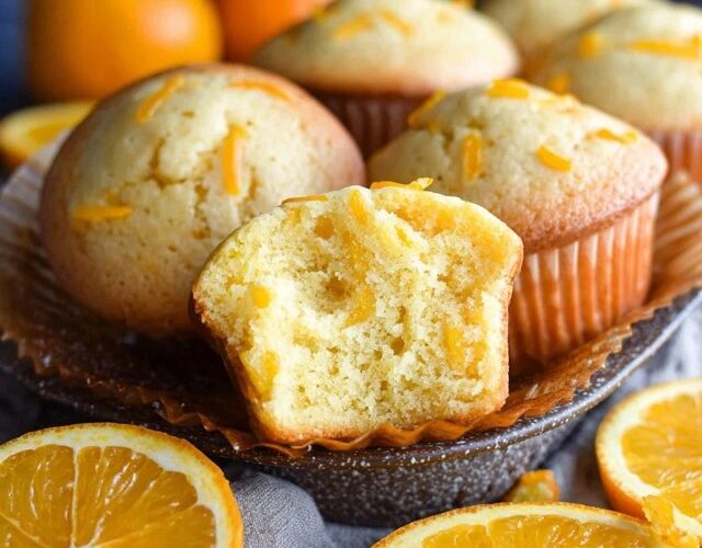 Orange muffins on a wire rack with golden domed tops, orange zest specks visible throughout, soft tender crumb, parchment liners, fresh oranges and zest curls around