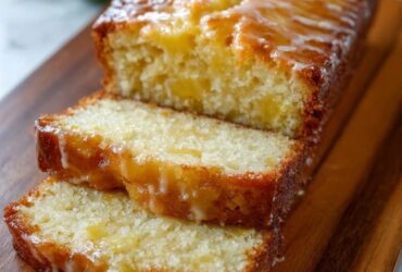 Lemon drizzle cake on a wooden board, sliced loaf showing tender yellow crumb, crackly sugar-lemon glaze top with crystallized texture, lemon slices and zest curls
