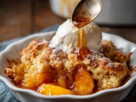 Peach cobbler in a white baking dish with golden buttery biscuit topping, juicy bubbling peach filling, scoop of vanilla ice cream melting on top, cinnamon dusting