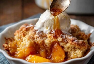 Peach cobbler in a white baking dish with golden buttery biscuit topping, juicy bubbling peach filling, scoop of vanilla ice cream melting on top, cinnamon dusting