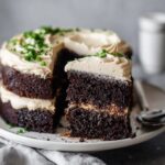 Chocolate layer cake on a white pedestal with thick swirls of vanilla buttercream frosting, dark moist chocolate layers visible, chocolate shavings on top, slice cut out