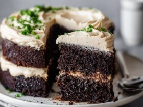 Chocolate layer cake on a white pedestal with thick swirls of vanilla buttercream frosting, dark moist chocolate layers visible, chocolate shavings on top, slice cut out