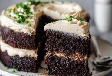 Chocolate layer cake on a white pedestal with thick swirls of vanilla buttercream frosting, dark moist chocolate layers visible, chocolate shavings on top, slice cut out