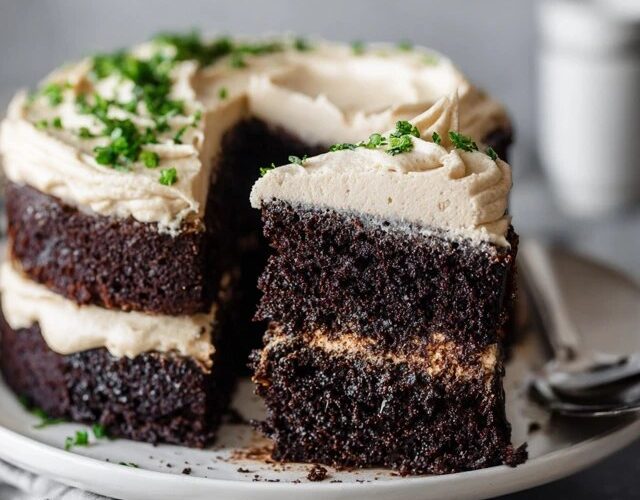 Chocolate layer cake on a white pedestal with thick swirls of vanilla buttercream frosting, dark moist chocolate layers visible, chocolate shavings on top, slice cut out