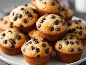 Chocolate chip muffins on a wire rack with tall domed golden tops, chocolate chips peeking through cracks, paper liners, soft tender crumb visible at edge