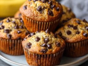 Chocolate chip banana muffins on a wooden board with golden domed tops, brown speckled banana surface, dark chocolate chips visible, ripe bananas in background