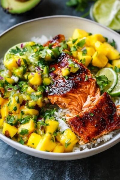 Mango salsa salmon bowl with crispy-skinned pan-seared salmon over white rice, bright orange mango salsa with red bell pepper, avocado slices, lime wedges, fresh cilantro