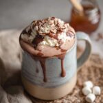 Two mugs of rich hot chocolate topped with towering swirls of fresh whipped cream, dusting of cocoa powder, chocolate shavings on top, marshmallows on the side, cozy wool blanket