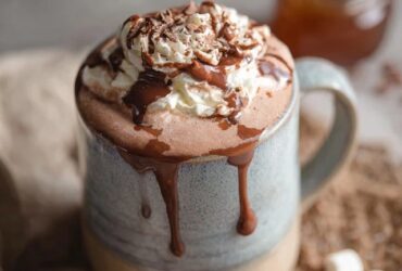 Two mugs of rich hot chocolate topped with towering swirls of fresh whipped cream, dusting of cocoa powder, chocolate shavings on top, marshmallows on the side, cozy wool blanket