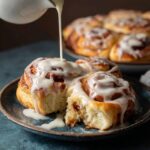 Cinnamon rolls with cream cheese icing in a baking dish, tall pillowy soft rolls with cinnamon-sugar swirls visible, thick white cream cheese glaze melting over warm tops, one roll lifted
