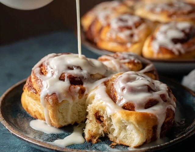 Cinnamon rolls with cream cheese icing in a baking dish, tall pillowy soft rolls with cinnamon-sugar swirls visible, thick white cream cheese glaze melting over warm tops, one roll lifted
