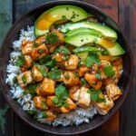 Teriyaki chicken rice bowl on a dark plate, glossy soy-glazed chicken thigh chunks over fluffy white rice, bright green steamed broccoli florets, toasted sesame seeds and sliced scallions on top, chopsticks resting beside
