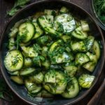 Cucumber dill salad in a white bowl, thin-sliced cucumber rounds tossed with creamy sour cream dressing, red onion slivers, plenty of fresh chopped dill, cracked black pepper, served on a wooden board