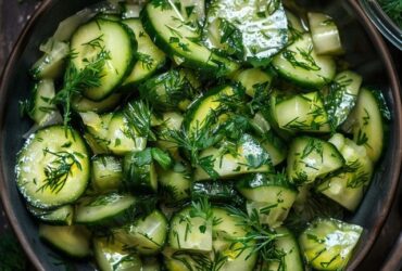 Cucumber dill salad in a white bowl, thin-sliced cucumber rounds tossed with creamy sour cream dressing, red onion slivers, plenty of fresh chopped dill, cracked black pepper, served on a wooden board