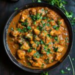 Chicken curry in a copper karahi pan, bone-in chicken pieces in deep orange-red tomato-onion gravy with visible spice flecks, cilantro garnish, basmati rice and naan on the side