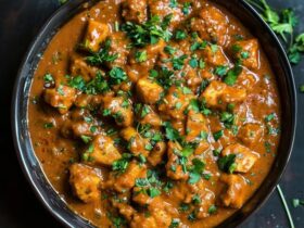 Chicken curry in a copper karahi pan, bone-in chicken pieces in deep orange-red tomato-onion gravy with visible spice flecks, cilantro garnish, basmati rice and naan on the side