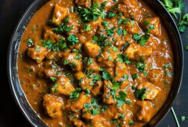 Chicken curry in a copper karahi pan, bone-in chicken pieces in deep orange-red tomato-onion gravy with visible spice flecks, cilantro garnish, basmati rice and naan on the side