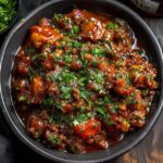 Vegetable manchurian gravy in a black bowl, golden-fried vegetable balls floating in glossy dark-red gravy with onion bell pepper specks, scallion garnish, fried rice in a small bowl beside