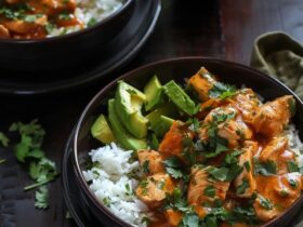 Chicken rice bowl with avocado on a wooden board, golden seared chicken cubes over fluffy white rice, half a sliced ripe avocado fanned across the top, fresh cilantro, lime wedges, drizzle of chipotle crema