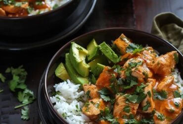 Chicken rice bowl with avocado on a wooden board, golden seared chicken cubes over fluffy white rice, half a sliced ripe avocado fanned across the top, fresh cilantro, lime wedges, drizzle of chipotle crema