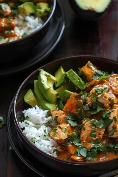 Chicken rice bowl with avocado on a wooden board, golden seared chicken cubes over fluffy white rice, half a sliced ripe avocado fanned across the top, fresh cilantro, lime wedges, drizzle of chipotle crema