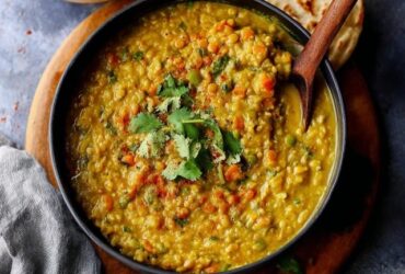 Lentil vegetable curry in a ceramic bowl, golden-orange red lentil base with chunks of carrot, wilted spinach, diced tomato, swirl of coconut milk on top, fresh cilantro garnish, basmati rice and naan on the side