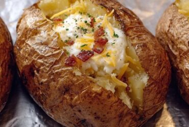 Loaded baked potato split open on a white plate, fluffy white potato interior topped with melted cheddar cheese, crispy crumbled bacon, sour cream dollop, sliced green chives, pat of butter melting, crispy salted skin