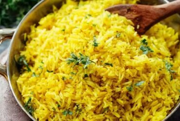 Turmeric rice with herbs in a white serving bowl, vibrant golden-yellow fluffy basmati grains studded with green chopped parsley and cilantro, cumin seed specks, sliced almonds and toasted pine nuts on top, a wedge of lemon on the side