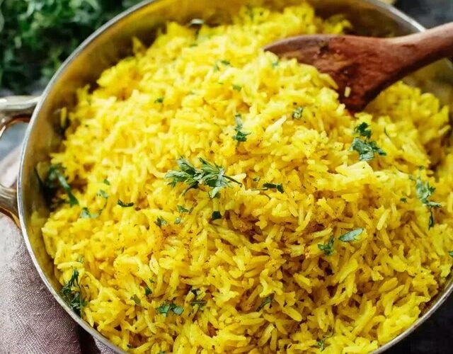 Turmeric rice with herbs in a white serving bowl, vibrant golden-yellow fluffy basmati grains studded with green chopped parsley and cilantro, cumin seed specks, sliced almonds and toasted pine nuts on top, a wedge of lemon on the side
