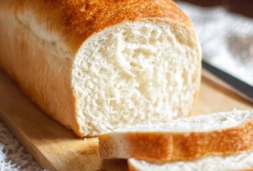 White sandwich bread sliced on a wooden cutting board, golden-brown crust with pillowy soft white interior, even uniform crumb structure visible in cut slices, butter dish and knife beside, kitchen towel underneath