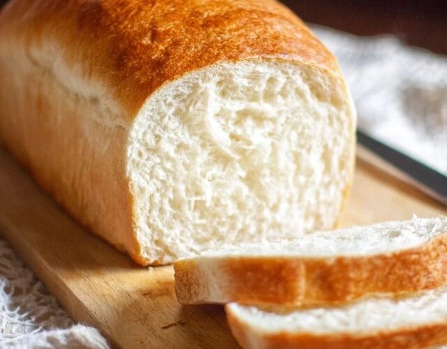 White sandwich bread sliced on a wooden cutting board, golden-brown crust with pillowy soft white interior, even uniform crumb structure visible in cut slices, butter dish and knife beside, kitchen towel underneath