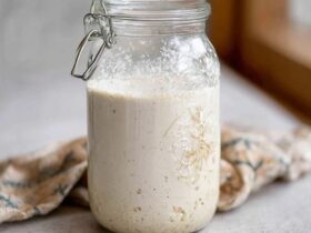 Sourdough starter in a clear glass jar on a kitchen counter, bubbly active starter doubling in size with visible air pockets, rubber band marking original level, wooden spoon and bag of flour beside, soft natural light from window