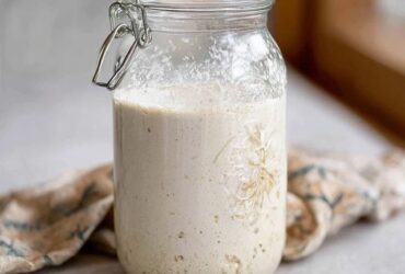 Sourdough starter in a clear glass jar on a kitchen counter, bubbly active starter doubling in size with visible air pockets, rubber band marking original level, wooden spoon and bag of flour beside, soft natural light from window