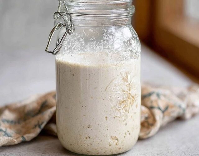 Sourdough starter in a clear glass jar on a kitchen counter, bubbly active starter doubling in size with visible air pockets, rubber band marking original level, wooden spoon and bag of flour beside, soft natural light from window